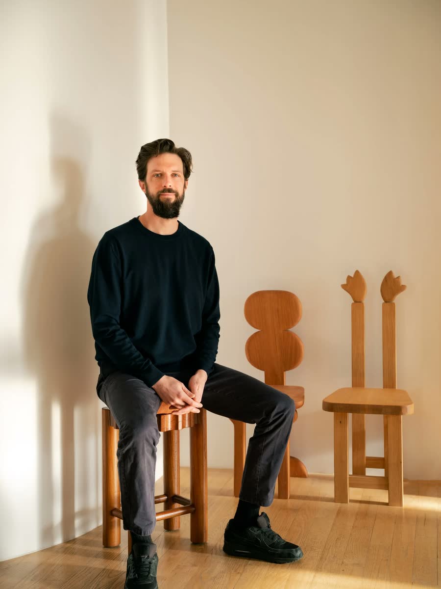 Oliver Haslegrave seated on a timber stool beside two sculptural chairs